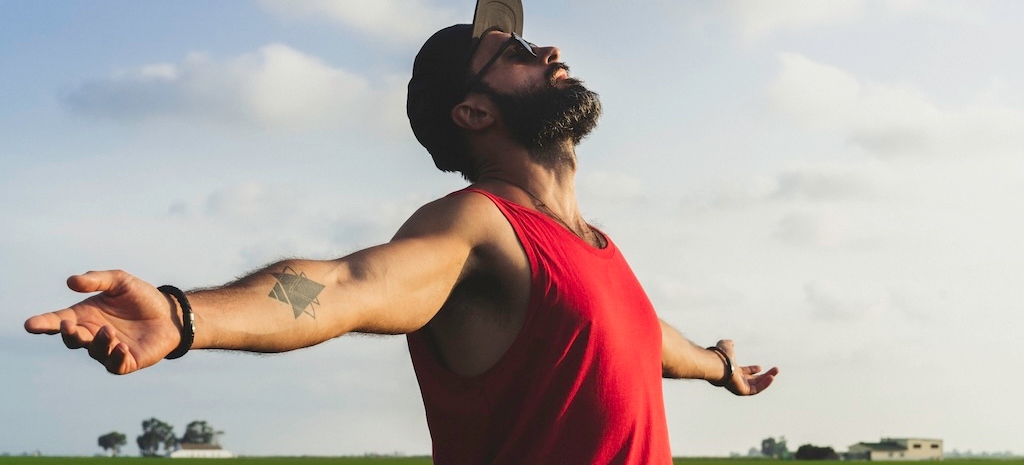 Bearded man model wearing black cap, sunglasses and red tshirt looks away, enjoing sunset.