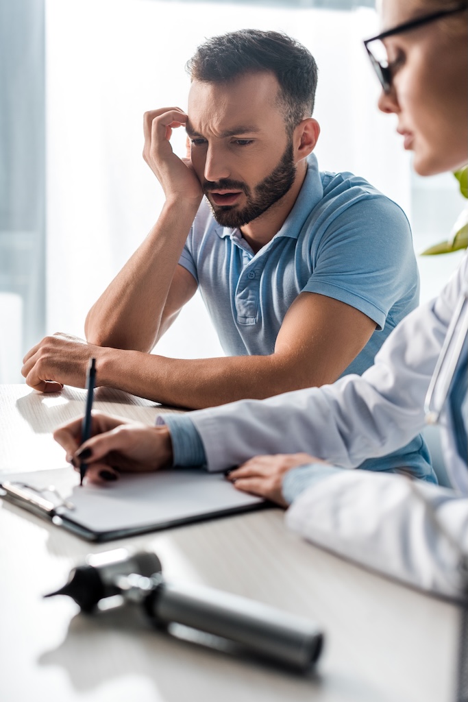 selective focus of bearded man looking at doctor in glasses writing diagnosis